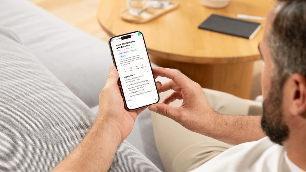 A man on a couch holding a phone that shows Mae’s Sheet Pan Chicken and Zucchini recipe card